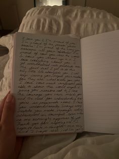 an open notebook with writing on it in someone's hand next to a bed