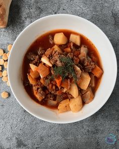 a white bowl filled with meat and vegetables on top of a gray surface next to bread