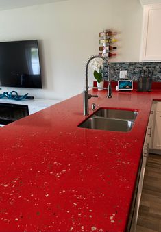 a red counter top in a kitchen next to a sink and microwave with a flat screen tv on the wall behind it