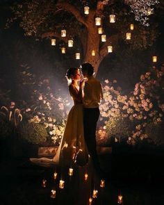 a man and woman standing under a tree with candles in the dark, surrounded by flowers