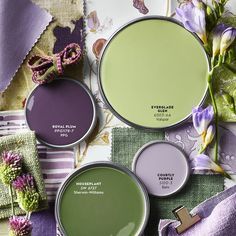 three different shades of green and purple paint with flowers on the table next to them