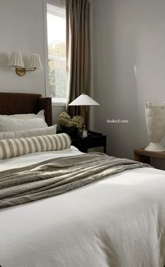 a bed with white sheets and pillows next to a lamp on a table in front of a window