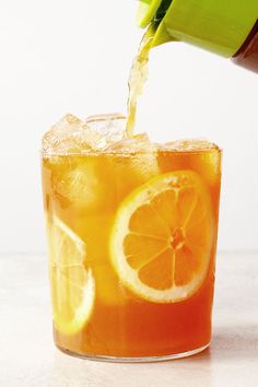 an orange drink being poured into a glass filled with ice and sliced lemon wedges