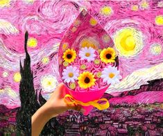 a hand holding a pink vase filled with sunflowers on top of a painting