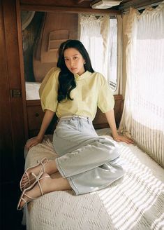 a young woman sitting on top of a bed next to a window with sheer curtains