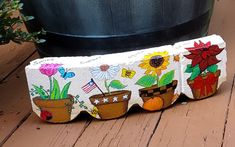 a painted rock with flowers and potted plants on it sitting next to a planter