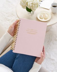 a woman is holding a pink notebook in her hands while sitting on a couch with white flowers