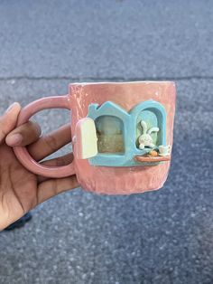 a hand holding a pink and blue coffee mug with a house on the inside, in front of a sidewalk