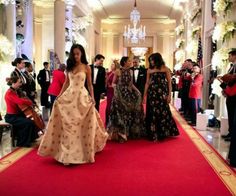 two women in dresses walking down a red carpeted hallway with other people standing around