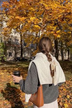 a woman standing in front of a tree with leaves on the ground and holding a coffee cup