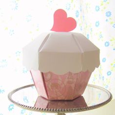 a paper cupcake with a heart on top sitting on a cake stand in front of a floral wallpaper