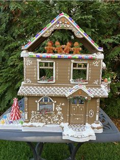 a gingerbread house on a table in front of some trees and bushes with decorations