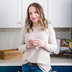 a woman standing in a kitchen holding a cup and looking at the camera with a smile on her face