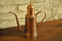 a copper colored coffee pot sitting on top of a wooden table next to a brick wall