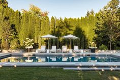 an outdoor pool with lounge chairs and umbrellas next to it in the middle of a lush green yard