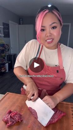 a woman with pink hair is cutting meat on a wooden table and smiling at the camera