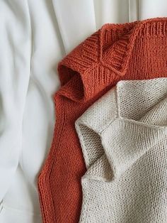 two sweaters laying next to each other on top of a white bed coverlet