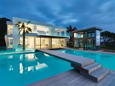 a large swimming pool in front of a modern house at night with palm trees on the deck