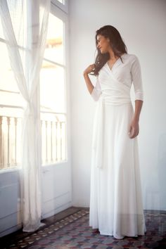 a woman standing in front of a window wearing a long white dress and holding her hand on her hip