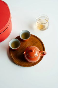 a tea pot and two cups on a wooden tray next to a red tin container