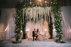 a bride and groom sitting on a couch in front of a floral covered stage at their wedding reception