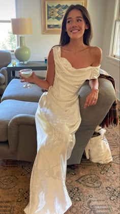 a woman in a white dress sitting on a couch holding a wine glass and smiling