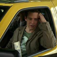 a young man sitting in the passenger seat of a yellow car holding his head with one hand