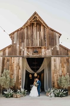 a bride and groom standing in front of a barn with lights strung from the roof