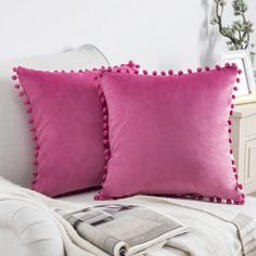 two pink pillows sitting on top of a white couch next to a magazine and potted plant