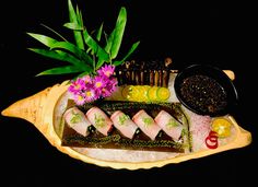 an assortment of sushi on a platter with dipping sauces and flowers in the background