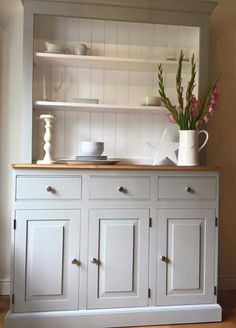 a white china cabinet with flowers in the vase and dishes on it's top