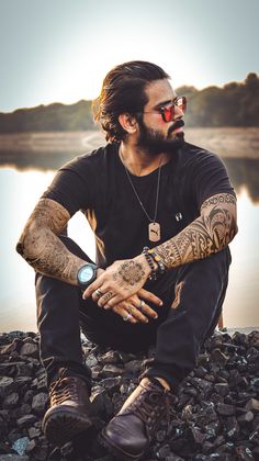 a man with tattoos sitting on rocks next to the water and holding his hands together