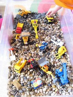 a plastic container filled with lots of toys and gravel next to a child's hand