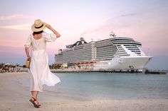 a woman in a white dress and hat walking on the beach with a cruise ship in the background