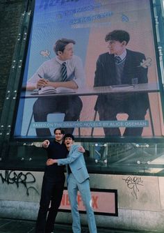 two men standing in front of a large poster on the side of a building with one holding his arm around another man's shoulder