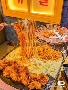 noodles being lifted from a pan with chopsticks over them in front of two plates of food