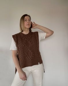 a woman standing in front of a white wall wearing a brown sweater and white pants