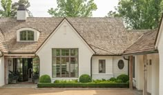 a white house with brown shingles on the roof and windows, surrounded by greenery