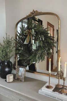 a mantle with a wreath and candles on it in front of a mirror that is reflecting the reflection of a house