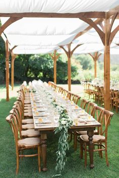 a long table set up with chairs and greenery for an outdoor wedding reception in the grass