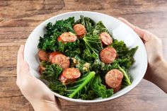 two hands holding a bowl filled with sausage and kale salad on top of a wooden table