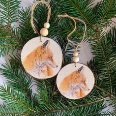 two wooden ornaments hanging from a tree with pine needles and white balls on them, one has an image of a red fox