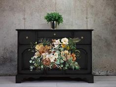 a black dresser with flowers painted on the front and side panels, next to a potted plant