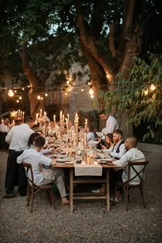 a group of people sitting at a table with candles in the middle of it and some trees behind them