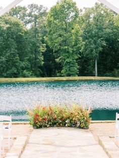 an outdoor ceremony with white chairs and flowers on the ground next to a body of water