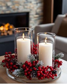 two candles are sitting on a glass plate with holly and red berries in front of a fireplace