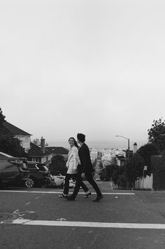 black and white photograph of two people walking across the street in front of parked cars