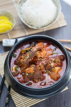 a bowl filled with meat and vegetables on top of a wooden table next to rice