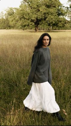 a woman standing in the middle of a field wearing a white skirt and gray sweater