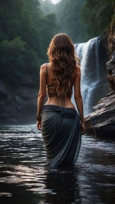 a woman standing in the water next to a waterfall with her back turned towards the camera